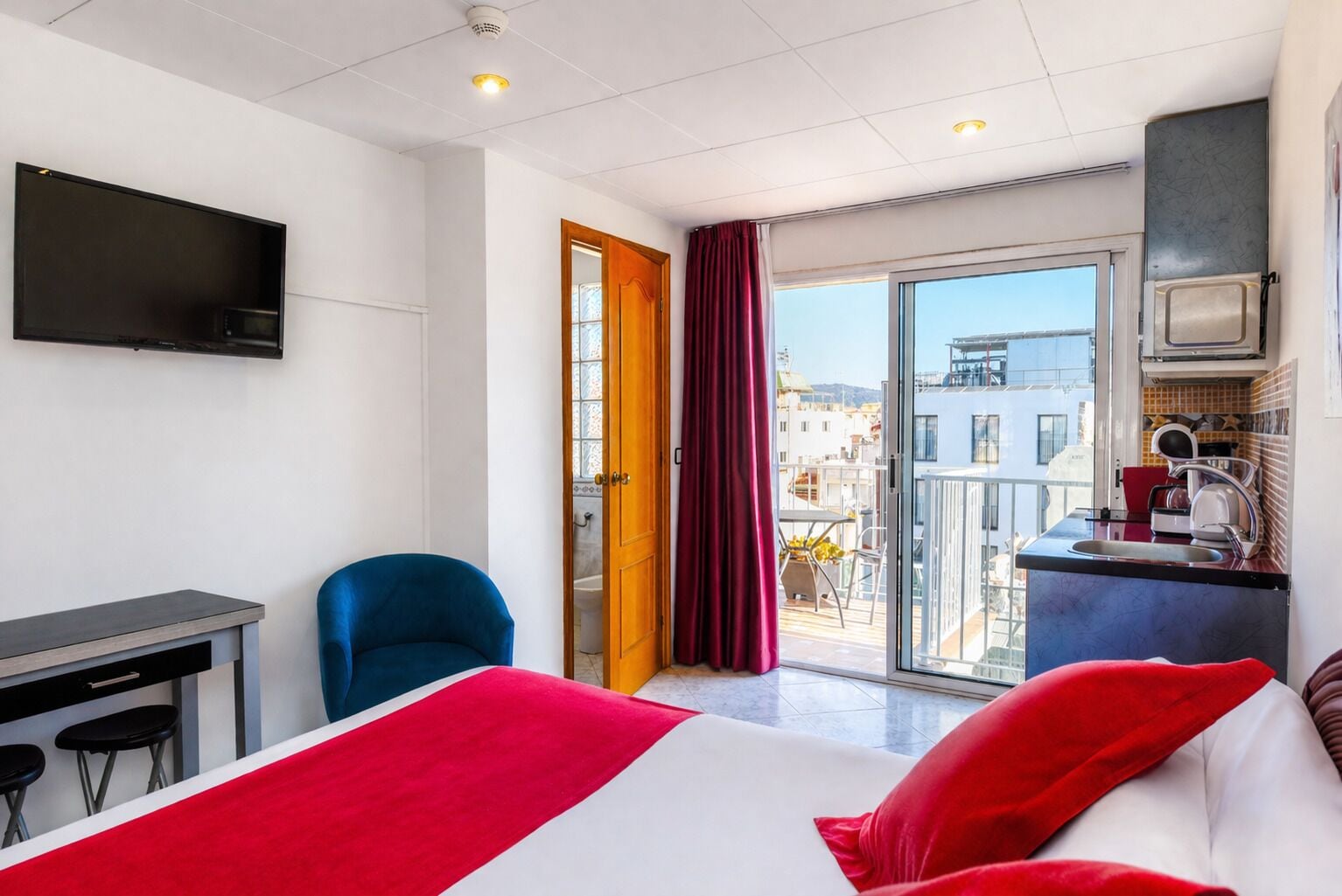 Habitación de hotel con cama, escritorio, silla, televisión, balcón con vistas y kitchenette.
