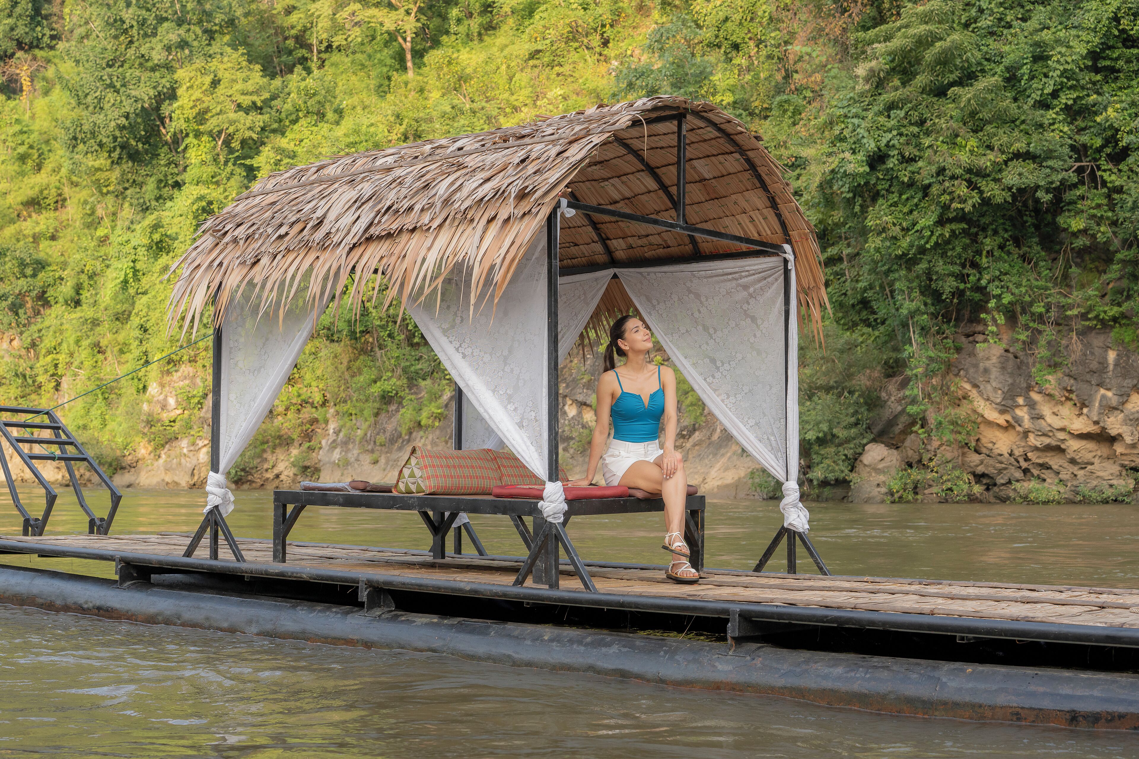 A thatched-roof structure with a person sitting inside, overlooking a body of water.