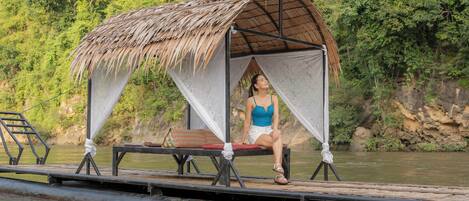 A thatched-roof structure with a person sitting inside, overlooking a body of water.