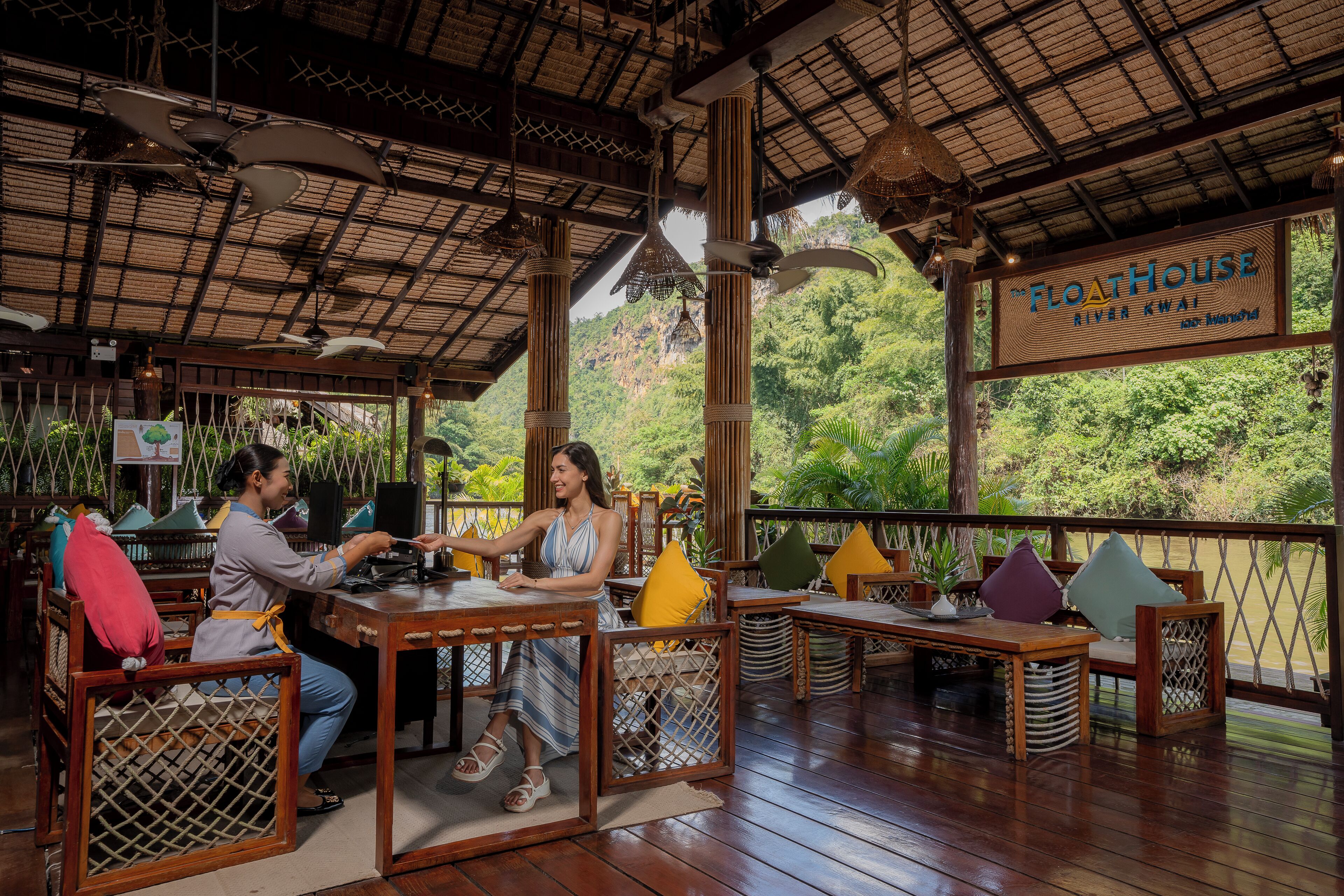 A wooden deck with tables and chairs, a sign reading "The Floathouse River Kwai", and a view of greenery and hills in the background.