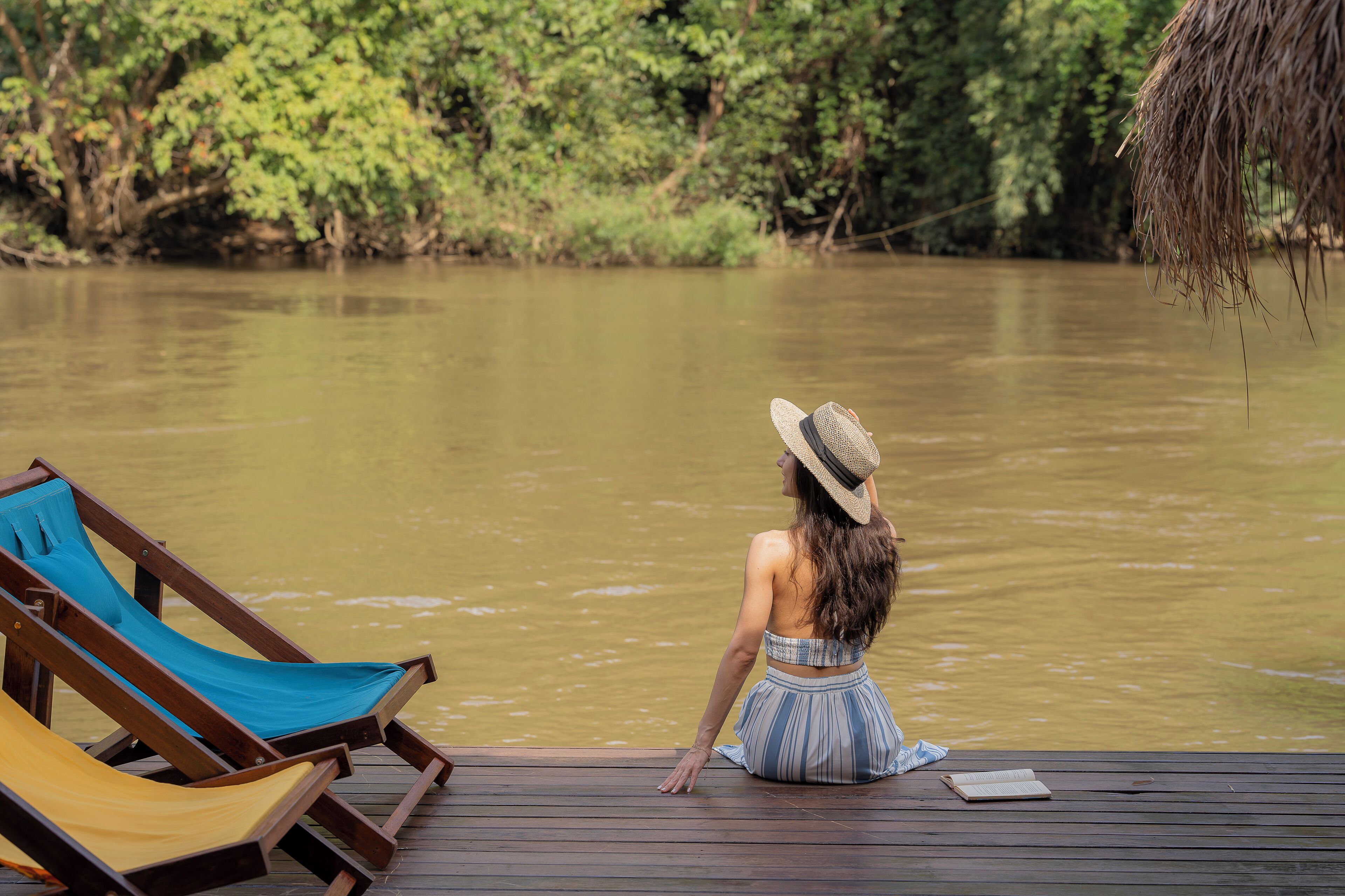 Een houten terras met lounge fauteuils en een persoon aan de rivier.