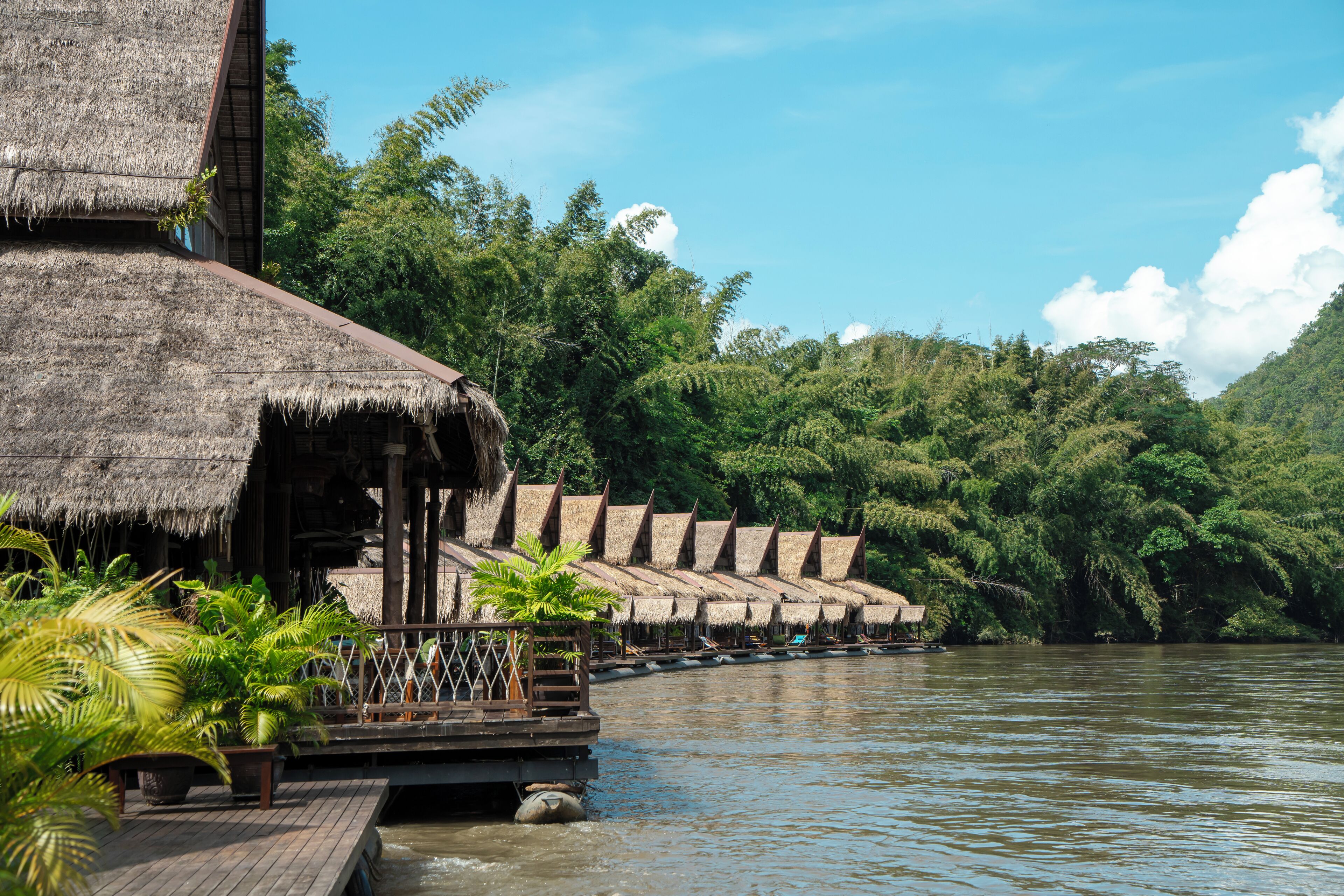 A thatched-roof structure with a wooden deck extending over a river, surrounded by lush greenery.
