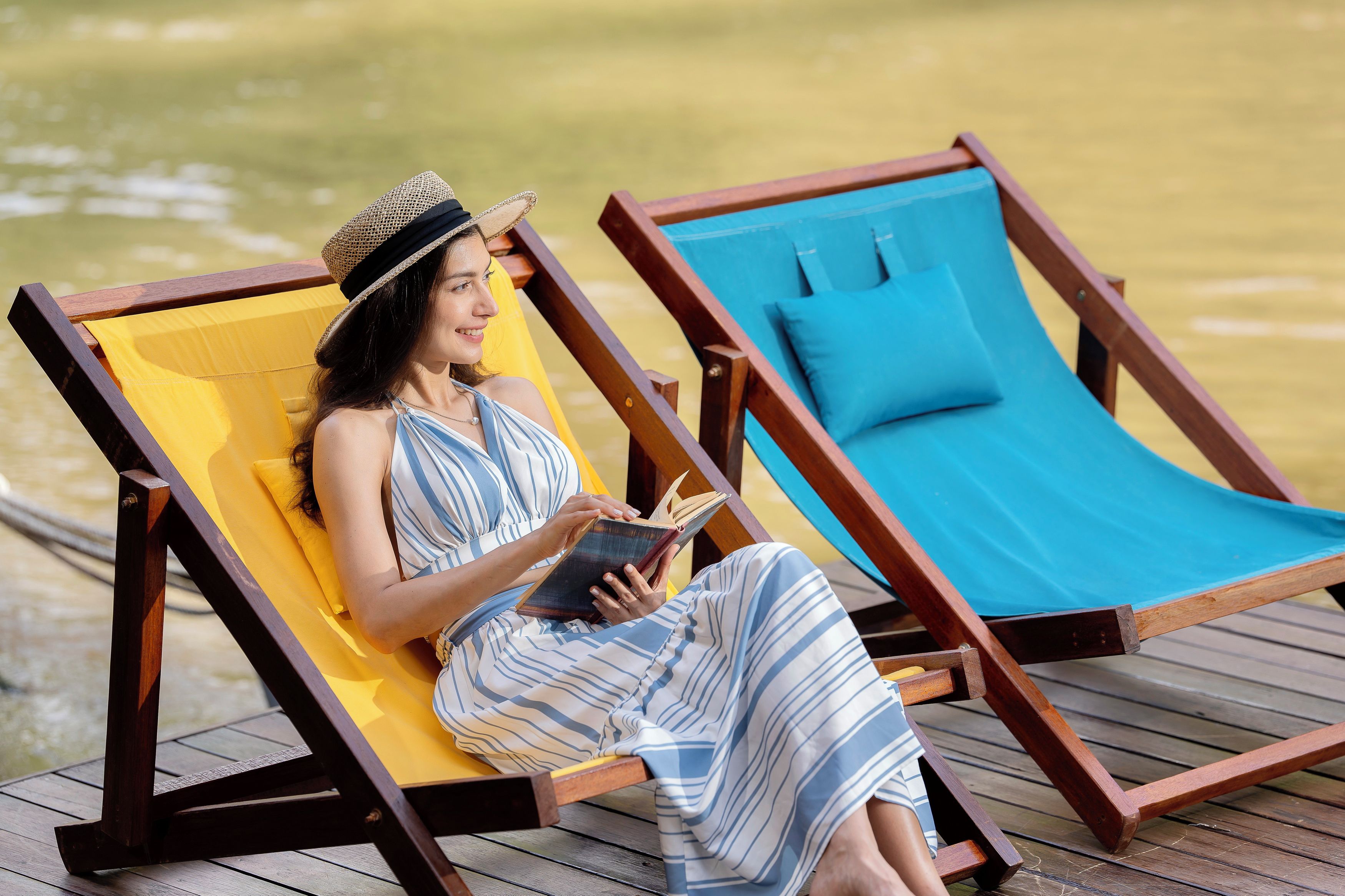 A person sitting on a wooden lounge chair with a yellow cushion, reading a book.
