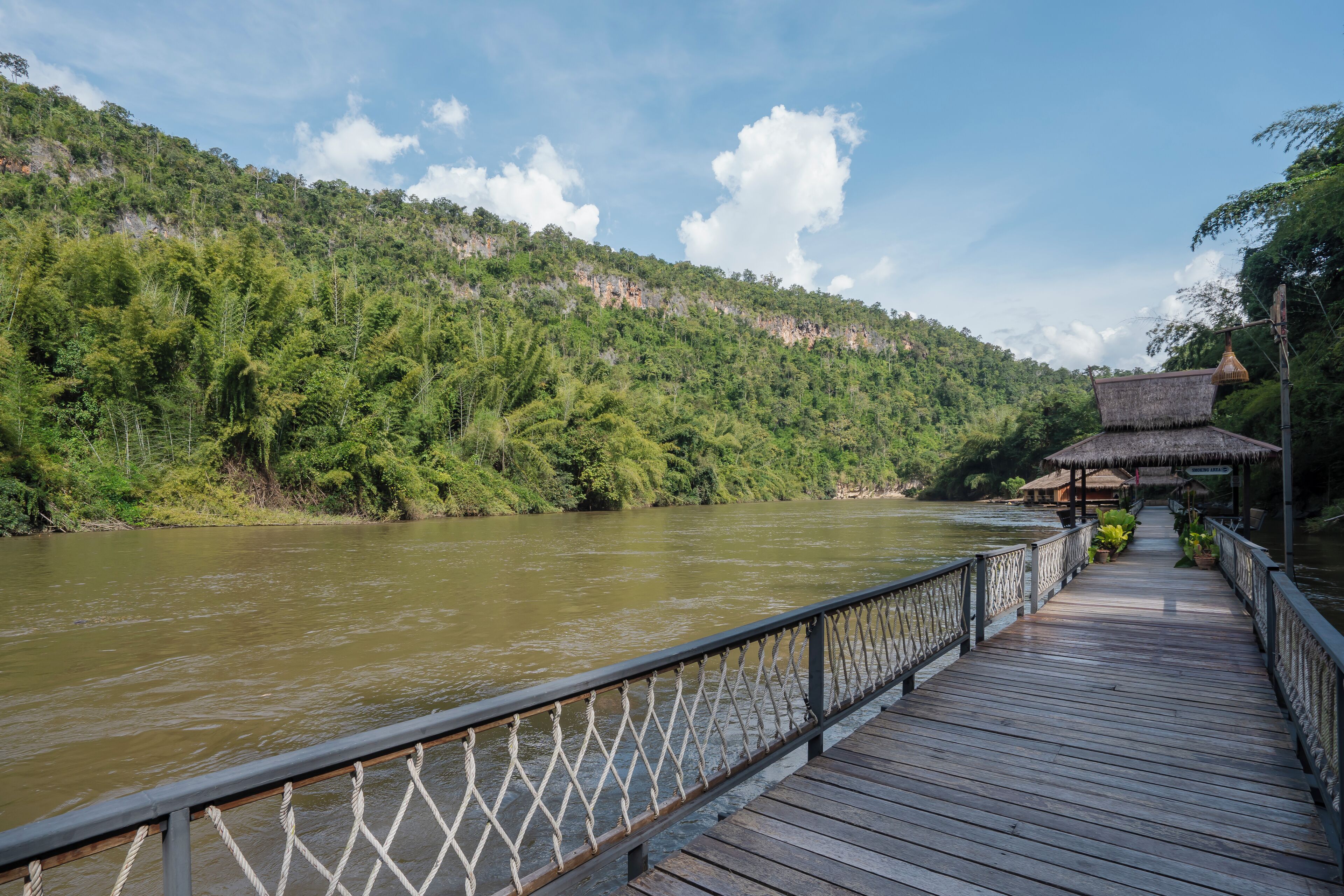 A wooden bridge with a railing leading to a thatched-roof structure by a river.