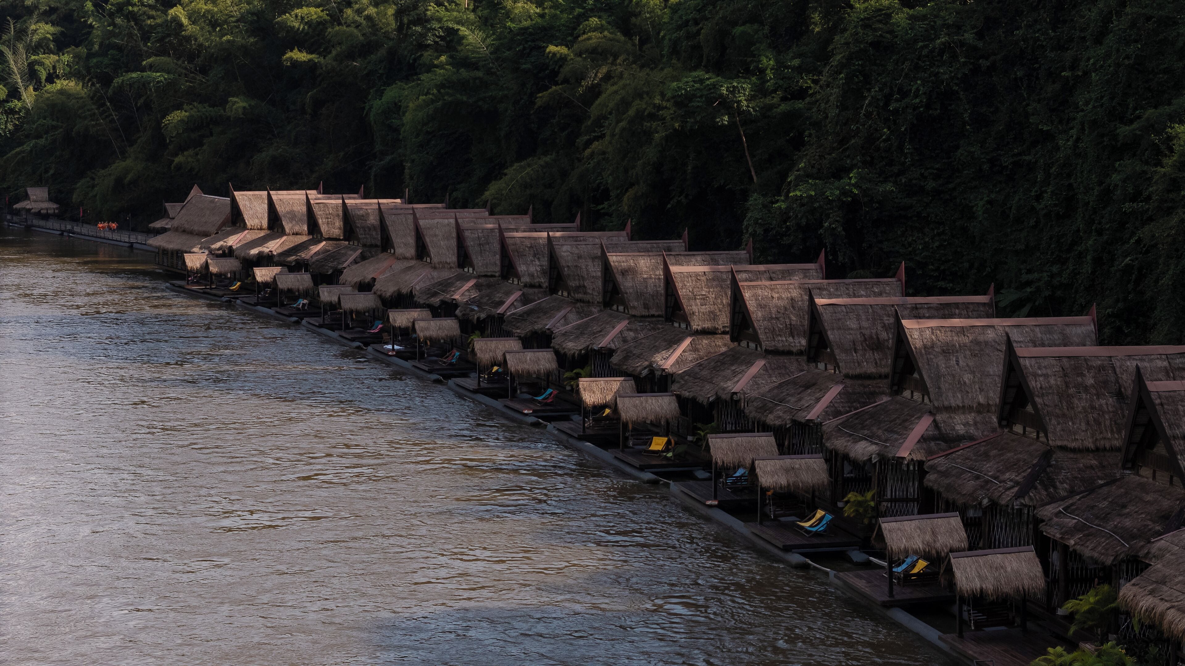 A row of thatched-roof huts along a riverbank, with a wooden walkway connecting them.