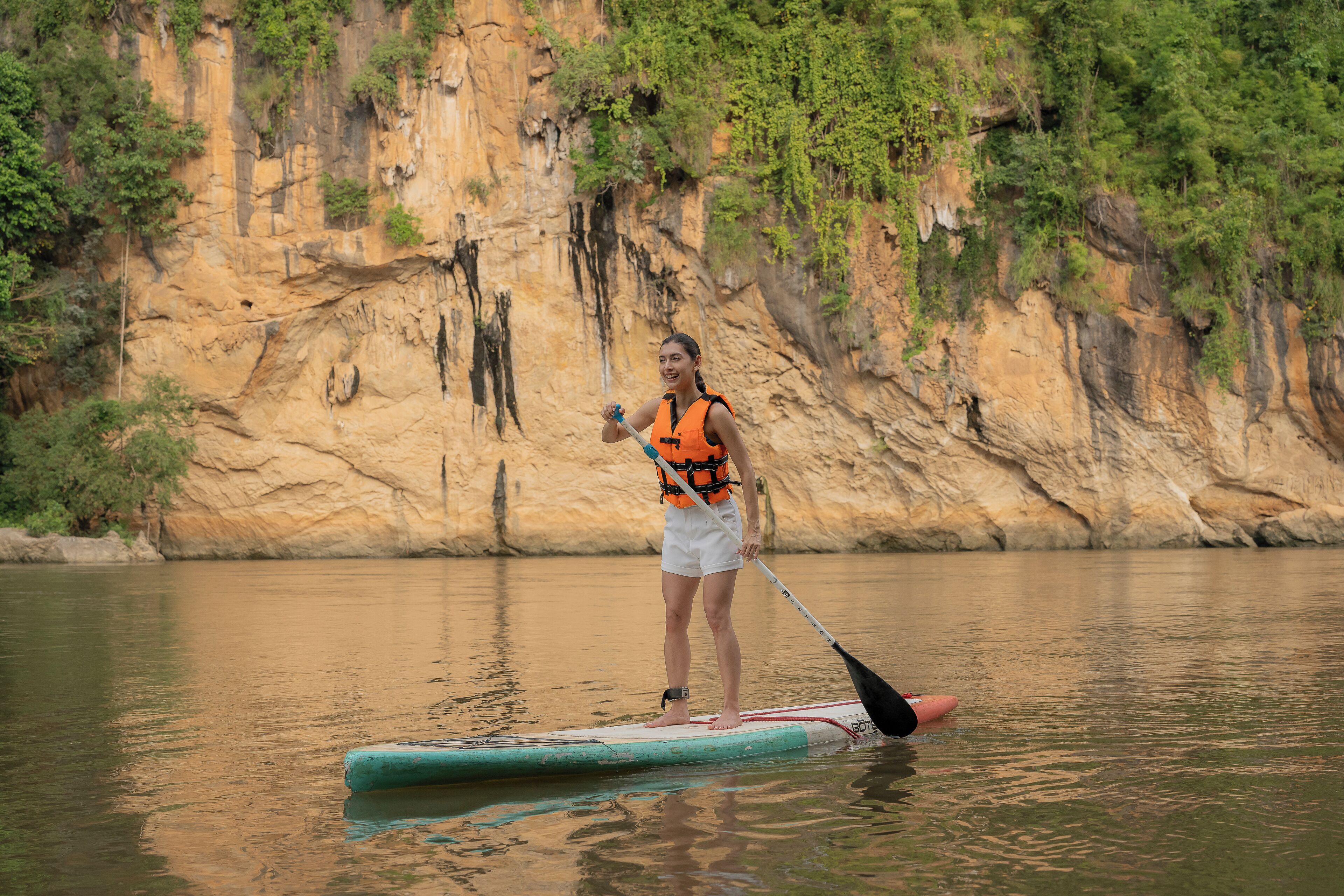 A person standing on a paddleboard in a calm body of water with a rocky cliff in the background.