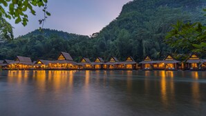 Una hilera de cabañas con techo de paja e iluminación cálida, con un denso bosque y un lago tranquilo de fondo.