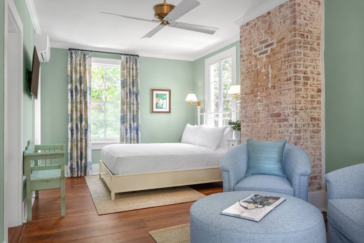 A bedroom with a bed, a chair, a desk, a ceiling fan, and a brick wall.