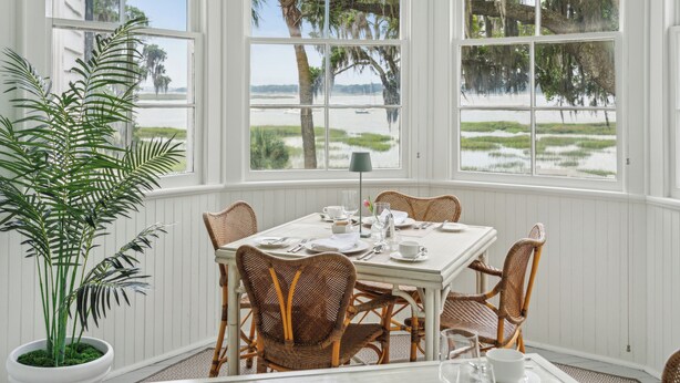 Un comedor con una mesa preparada para cuatro personas, rodeado de sillas de mimbre, ventanas amplias con vistas a una playa y humedales, y una planta en maceta.
