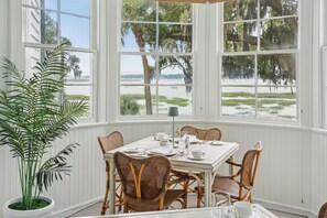 A dining area with a table set for four, surrounded by wicker chairs, large windows with a view of a beach and marshes, and a potted plant. - Cuthbert House (Beaufort)