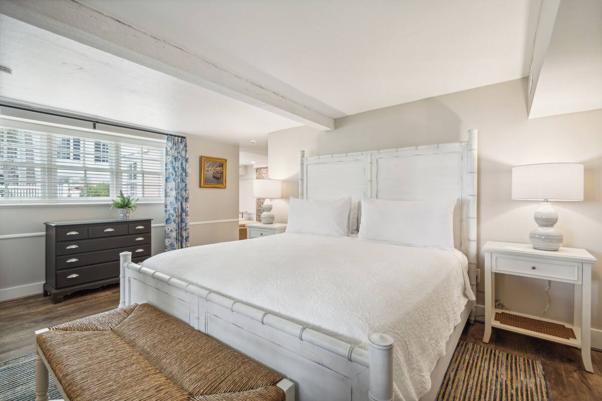 A bedroom with a large bed, a bedside table with a lamp, a dresser, and a window with blinds.