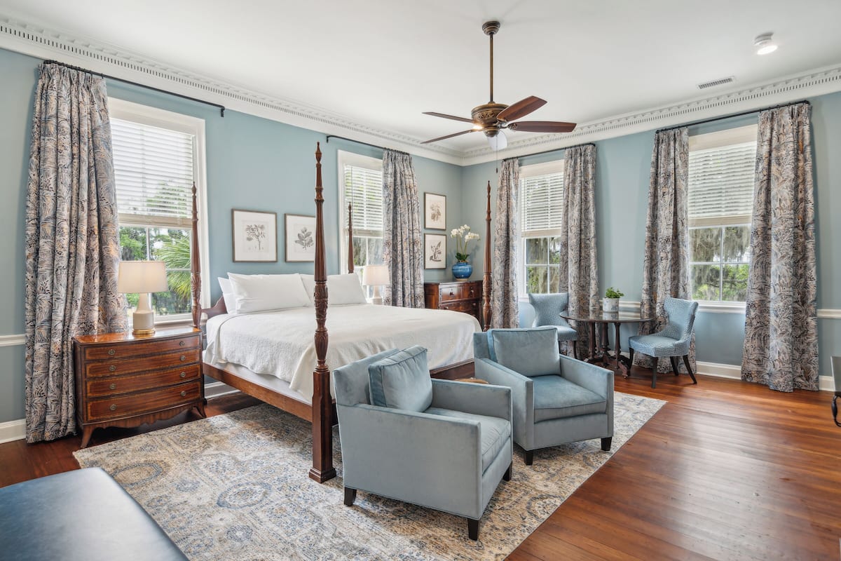 A bedroom with a four-poster bed, two armchairs, a nightstand, and a ceiling fan.