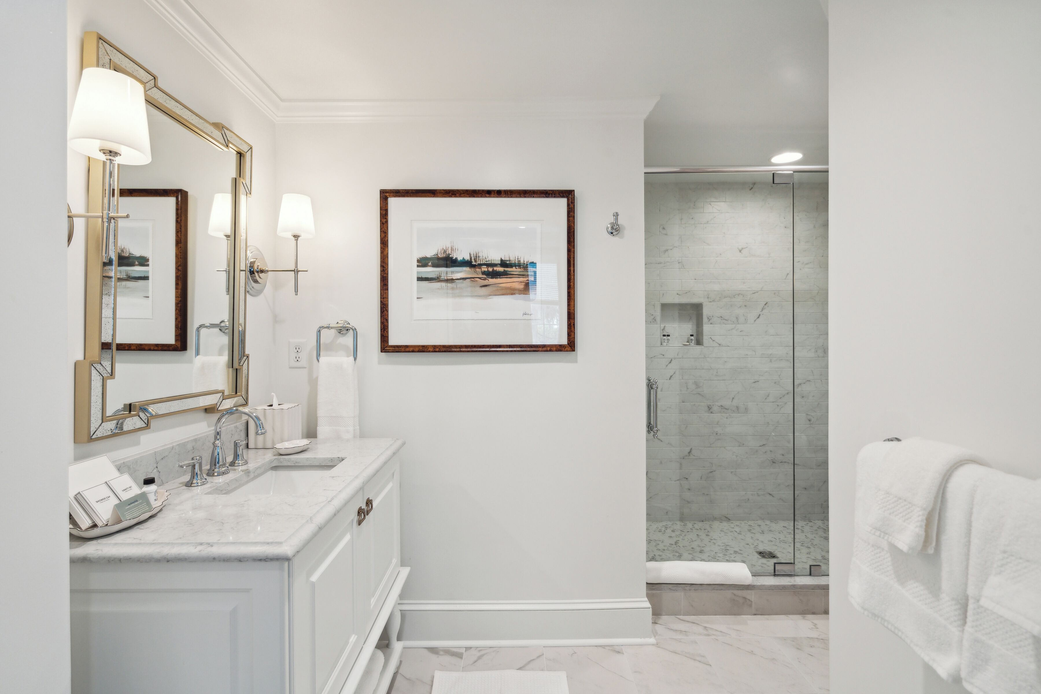 A modern bathroom with a large mirror, a framed picture, a marble sink, and a glass shower enclosure.