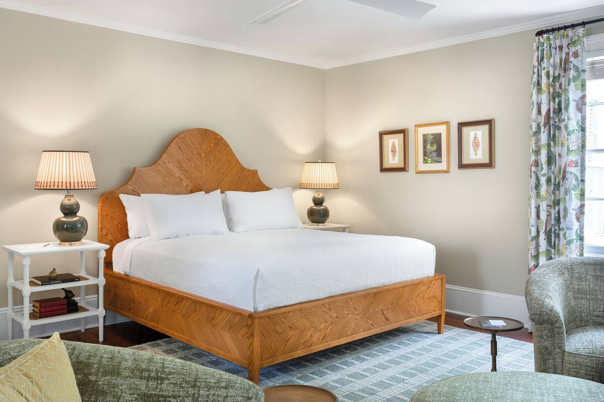 A neatly made bed with a wooden headboard, two bedside lamps, a small bedside table with books, and a chair in a room with a patterned rug.