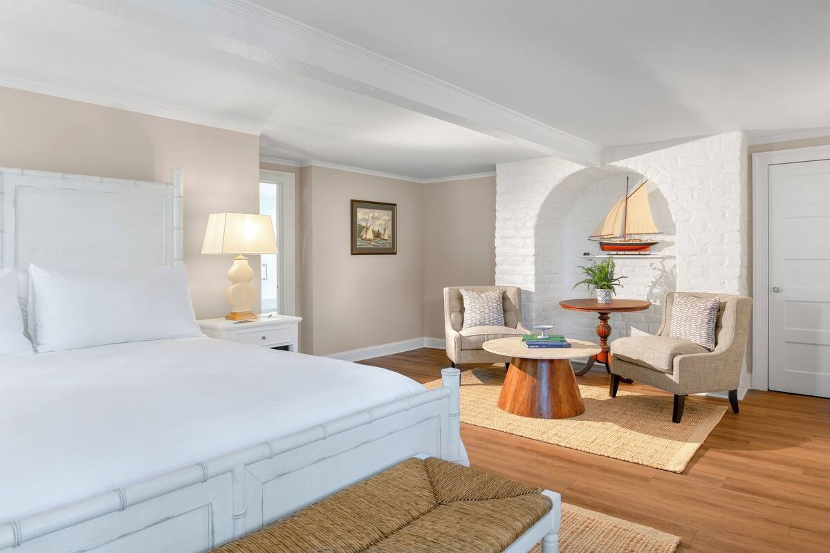 A bedroom with a large bed, two armchairs, a round coffee table, a lamp, and a small sailboat decoration.