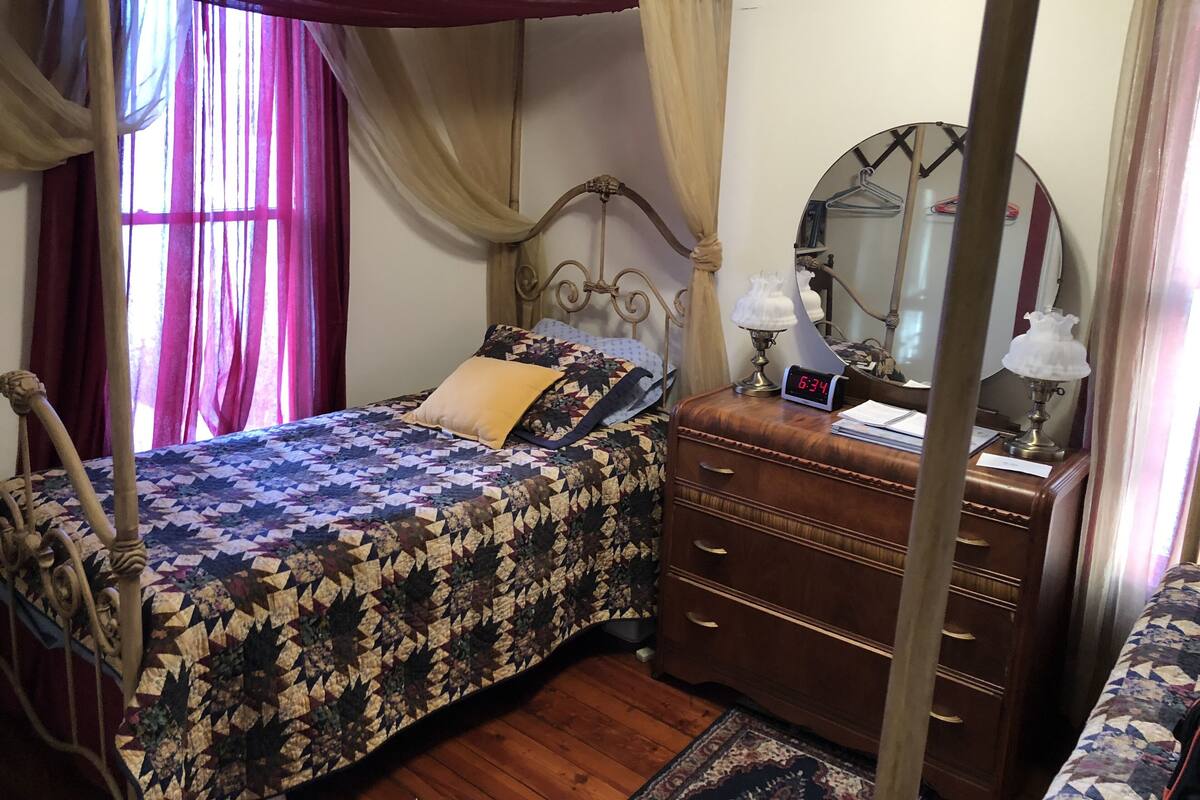 A four-poster bed with a patterned bedspread, a bedside table with a clock, and a mirror on the wall.