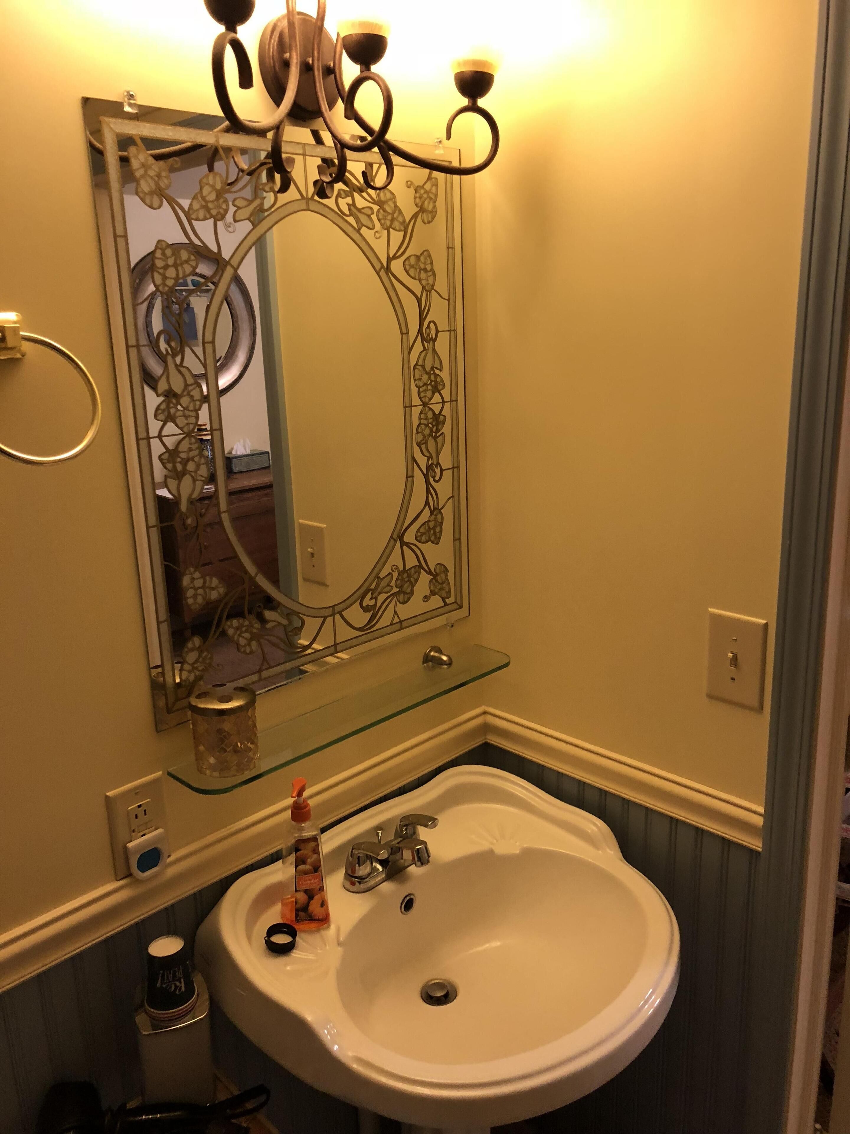 Un baño con un lavamanos blanco, un estante de vidrio arriba, un espejo decorativo con motivos florales y una lámpara de pared.
