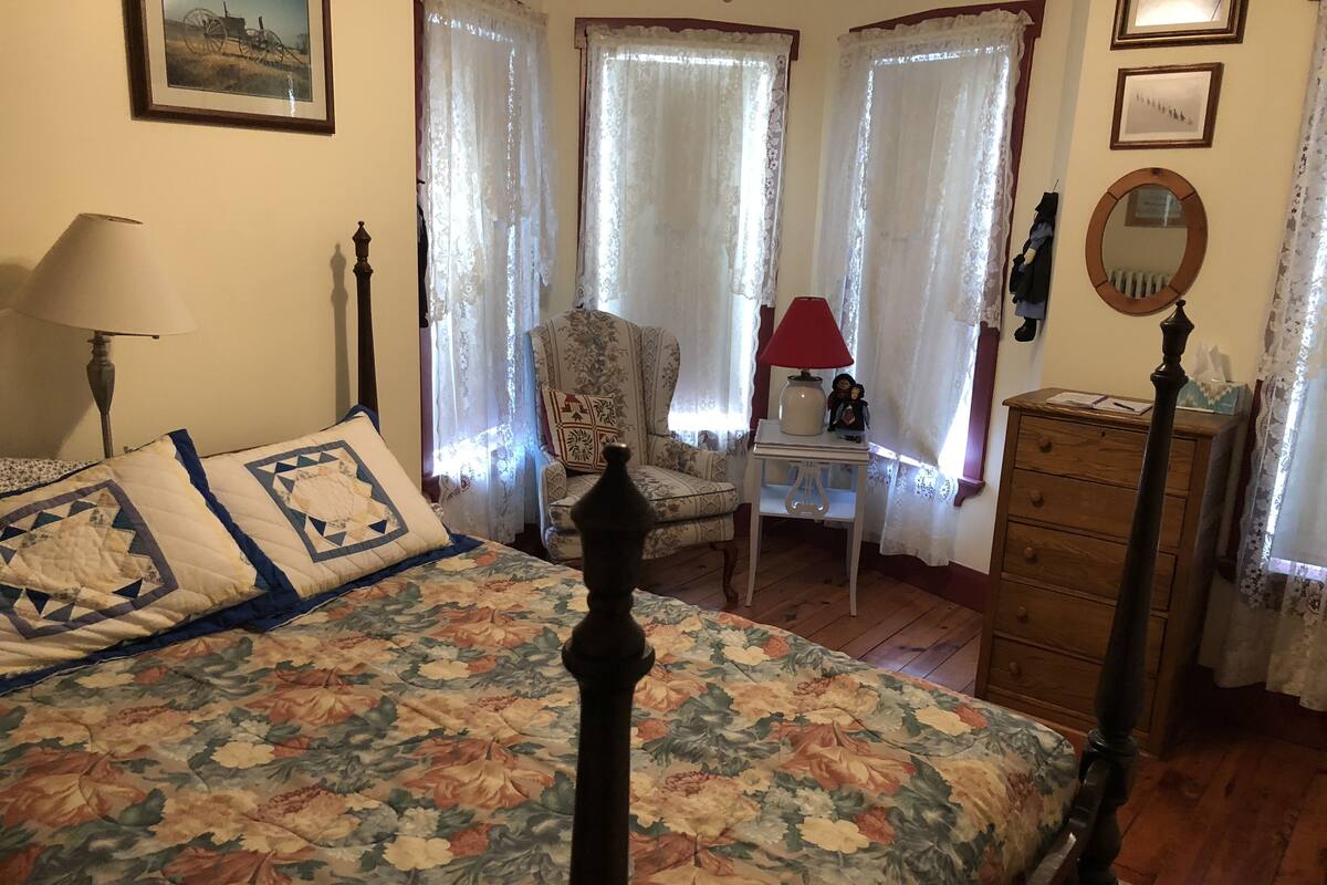 A bedroom with a floral bedspread, a wooden dresser, a chair, and a nightstand with a lamp.