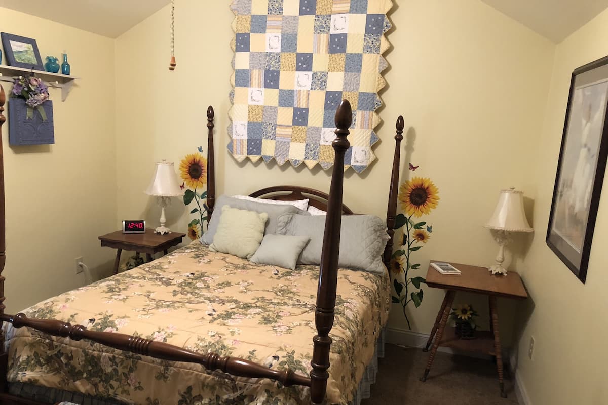 A bedroom with a four-poster bed, floral bedding, a quilt, a nightstand with a lamp, a wall clock, and a framed picture.