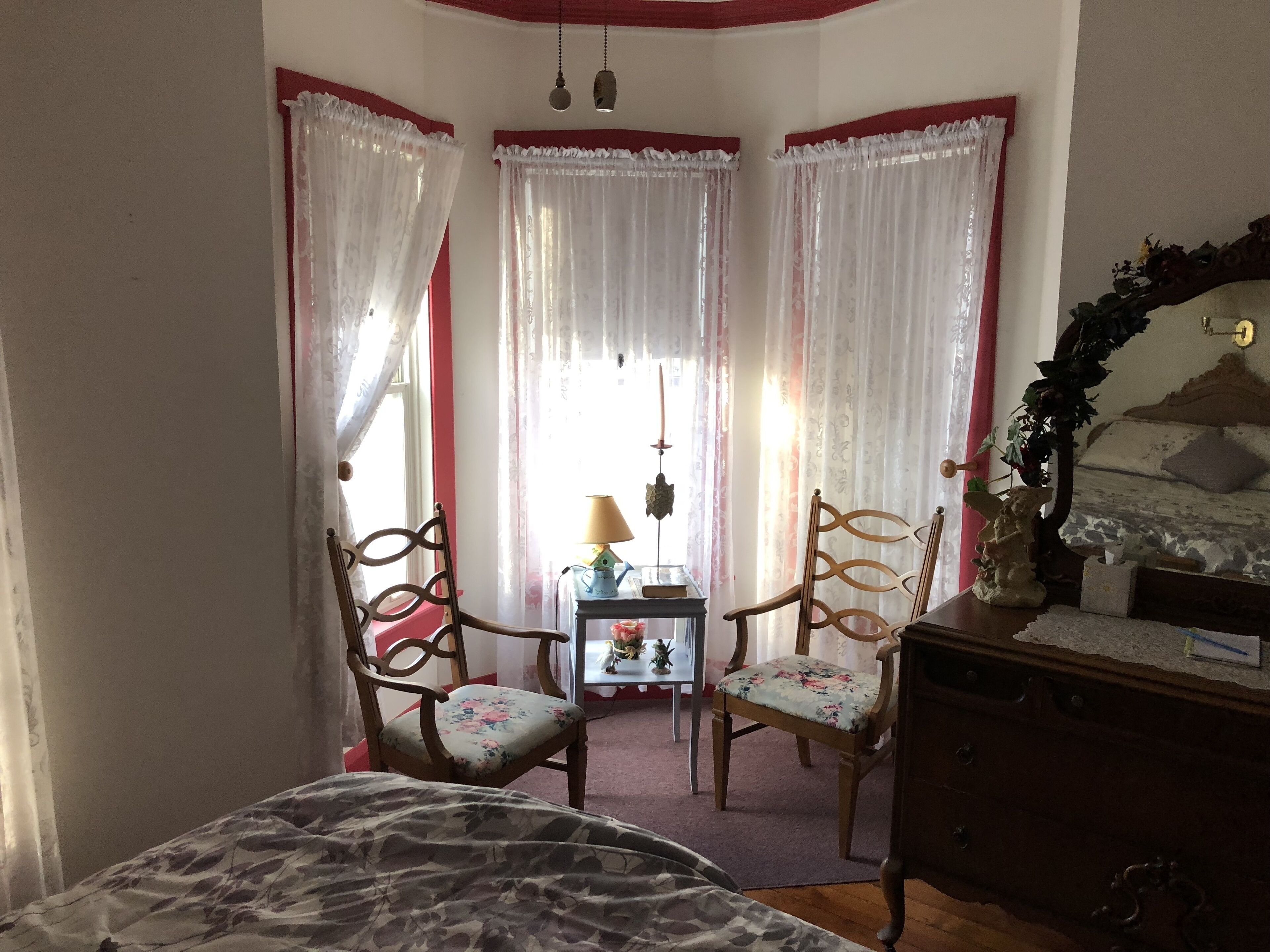 A bedroom with a bed, two chairs, a dresser, a small table with a lamp, and a window with curtains.