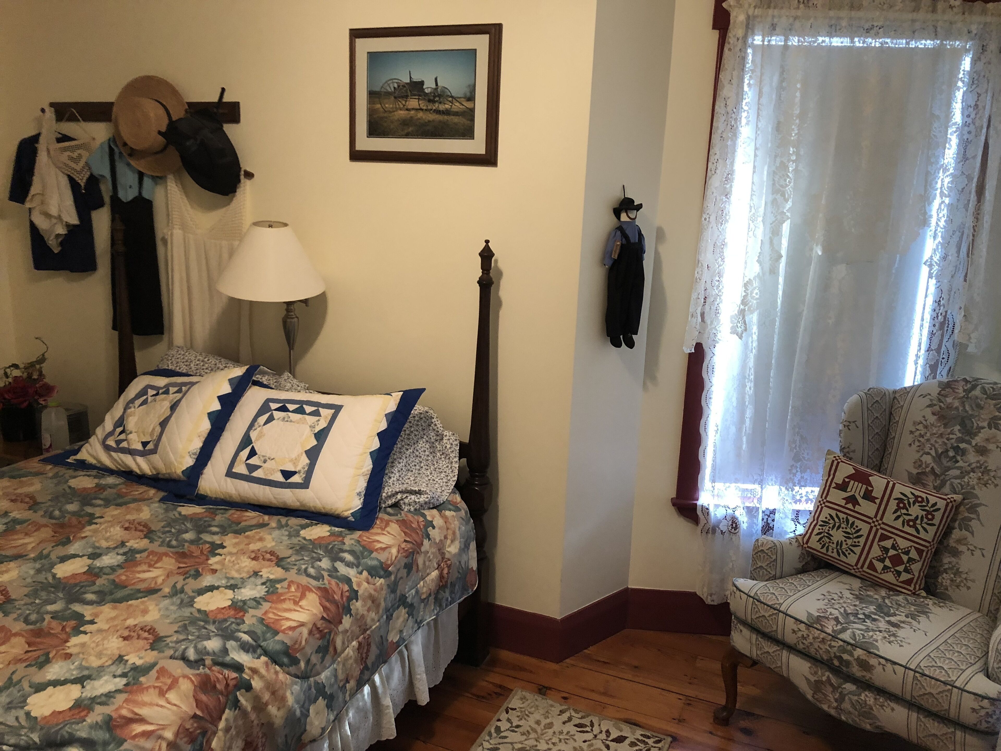 A bedroom with a floral bedspread, a wooden headboard, a nightstand with a lamp, a chair with patterned upholstery, and a window with sheer curtains.