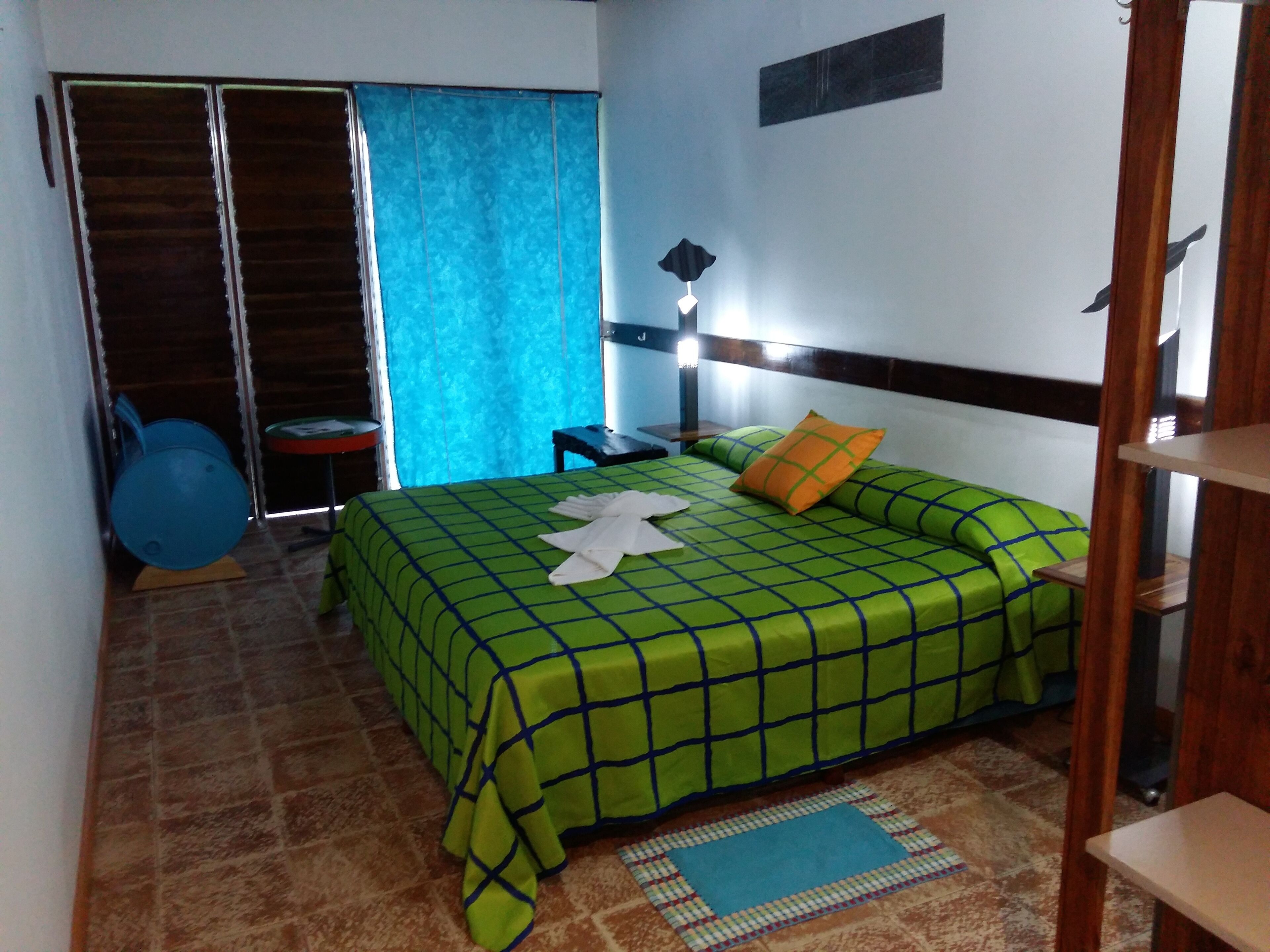 Un dormitorio con una colcha verde a cuadros, una cortina azul, un cabecero de madera, una mesita de noche y una silla azul.
