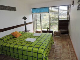 Un dormitorio con cama, mesita de noche, ventana con persianas, una pequeña mesa con una planta y vista a áreas verdes al exterior.