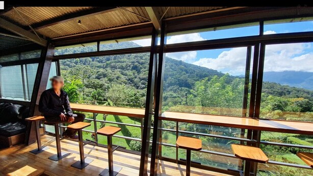 A room with large windows offering a view of a lush green landscape and mountains. There are wooden tables and chairs arranged for seating. - Celeste Mountain Lodge (Bijagua)