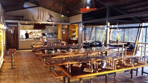 A restaurant with wooden tables and chairs, a kitchen area, and large windows. - Celeste Mountain Lodge (Bijagua)
