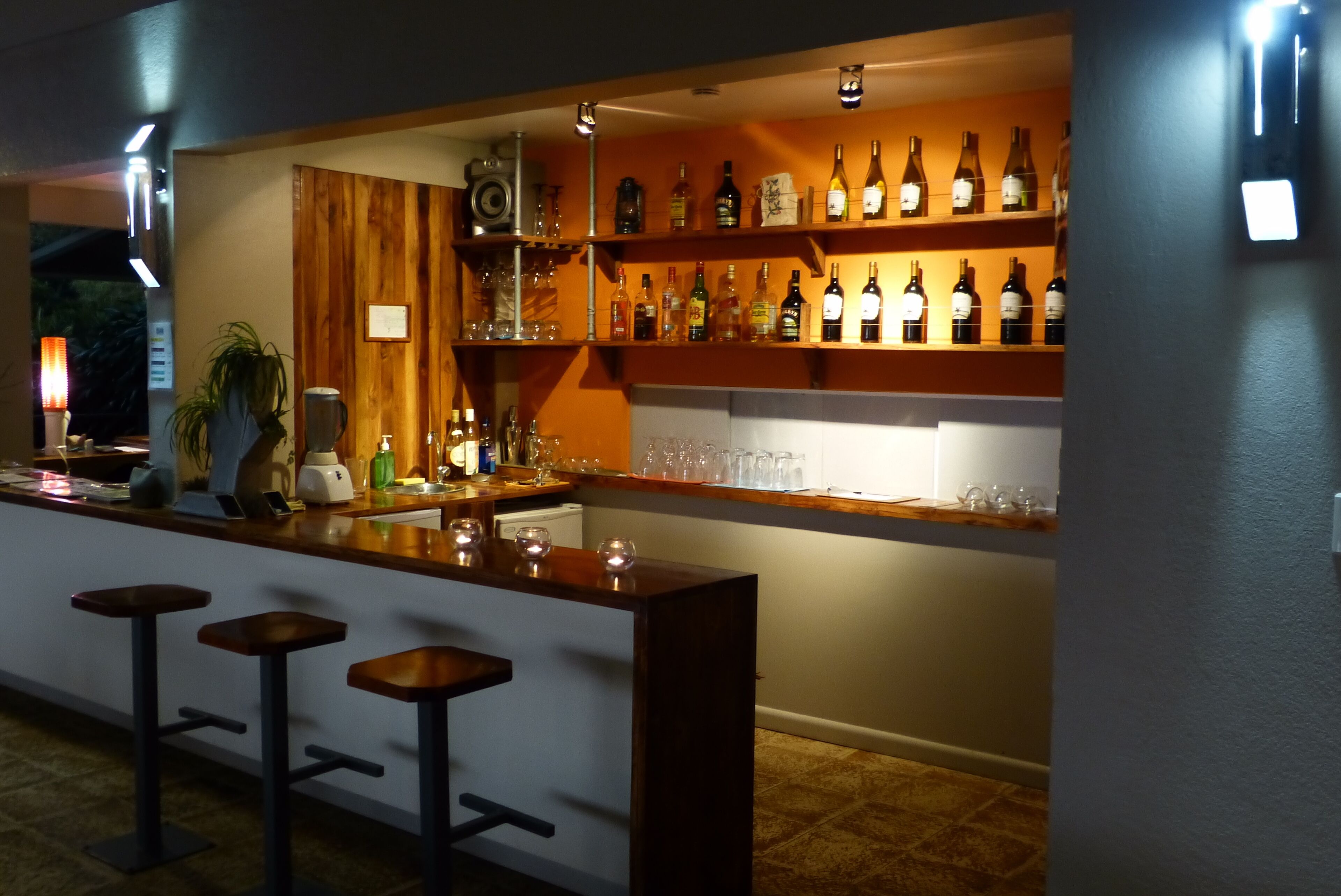 Un espace bar avec un comptoir en bois, des tabourets de bar et des étagères remplies de bouteilles et de verres.