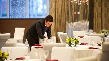 A person setting a table in a formal dining area with white chairs and tables, and a large window in the background.