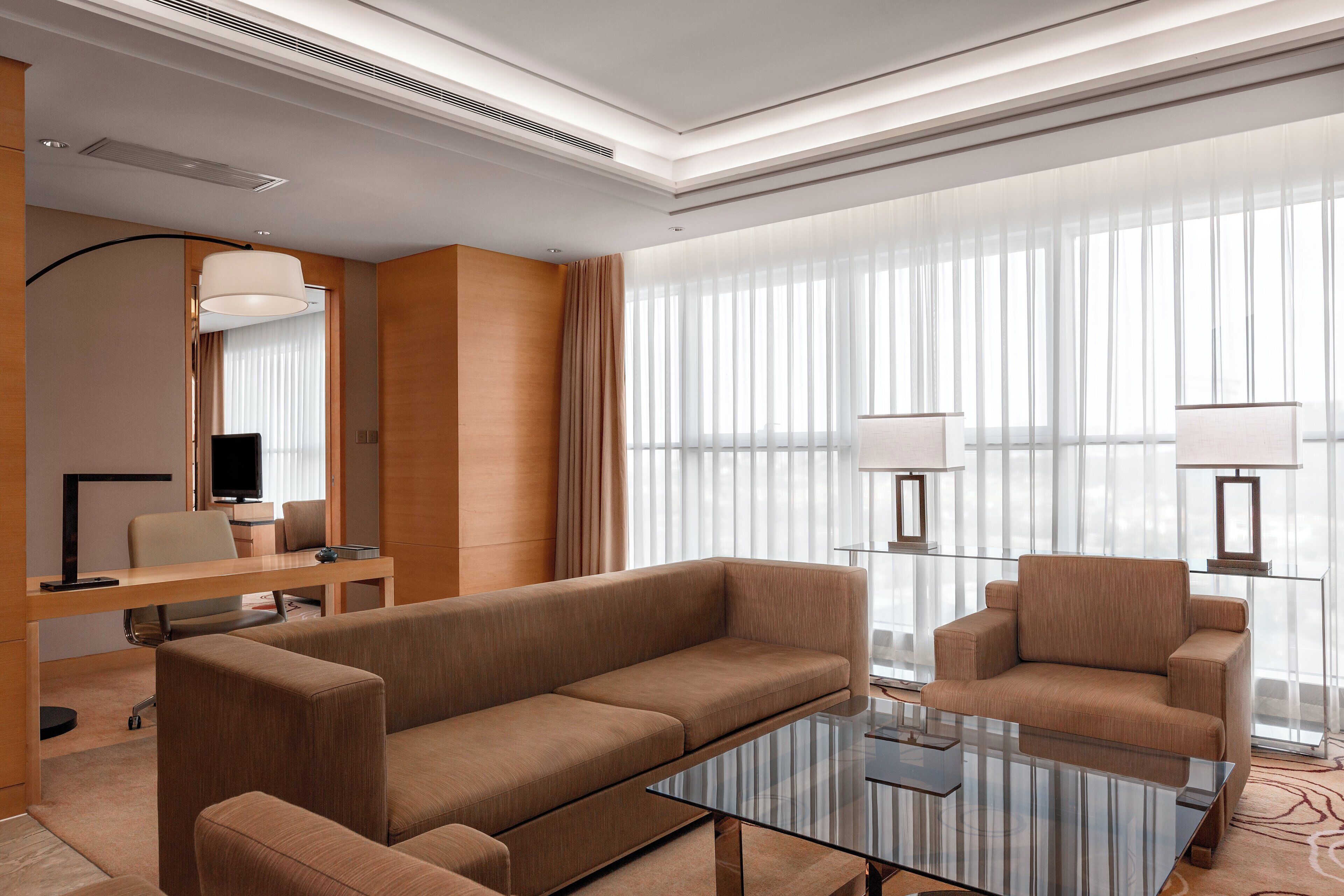 Una habitación de hotel moderna con un conjunto de sofá, una mesa de centro de vidrio, un televisor de pantalla plana y ventanas amplias con cortinas translúcidas.