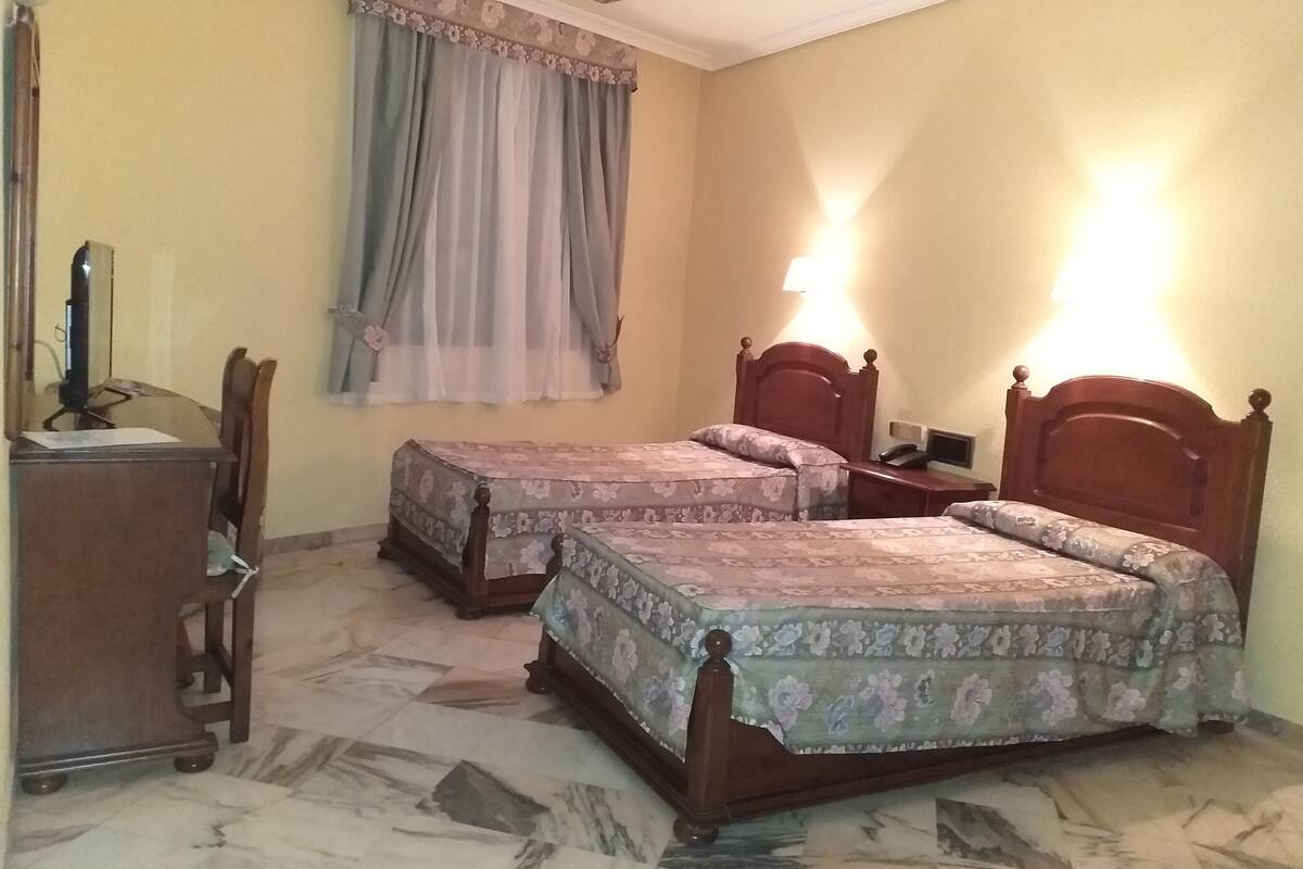 A hotel room with two beds, a desk with a chair, and a window with curtains.