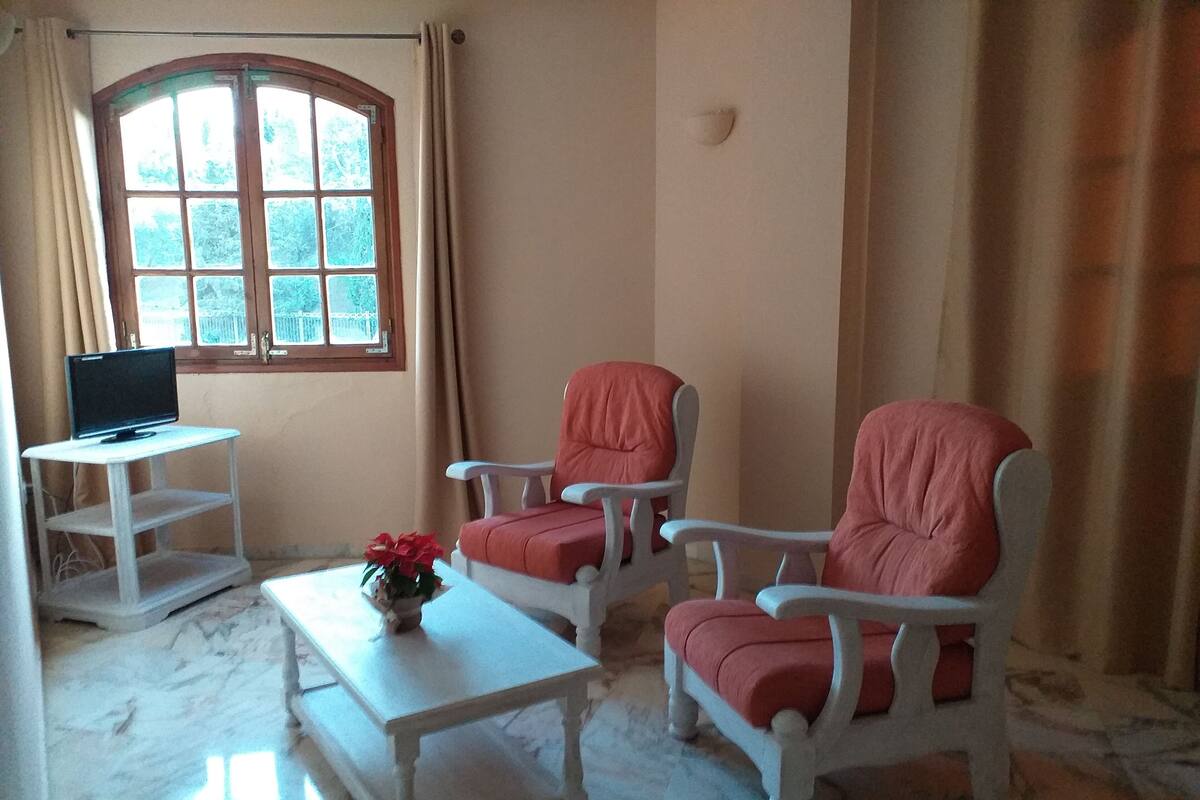 A room with two armchairs, a small table, a television on a stand, and a window with curtains.