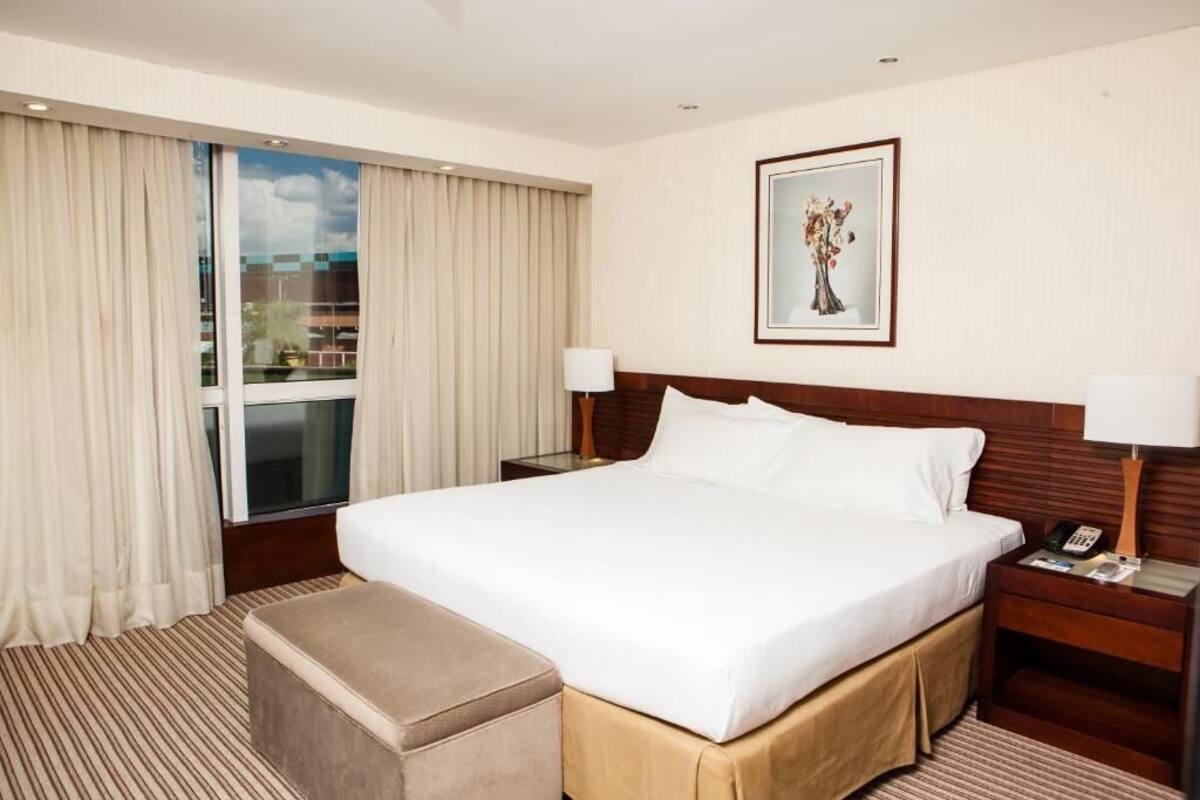 A hotel room with a large bed, a bedside table with a lamp, a window with curtains, and a framed picture on the wall.