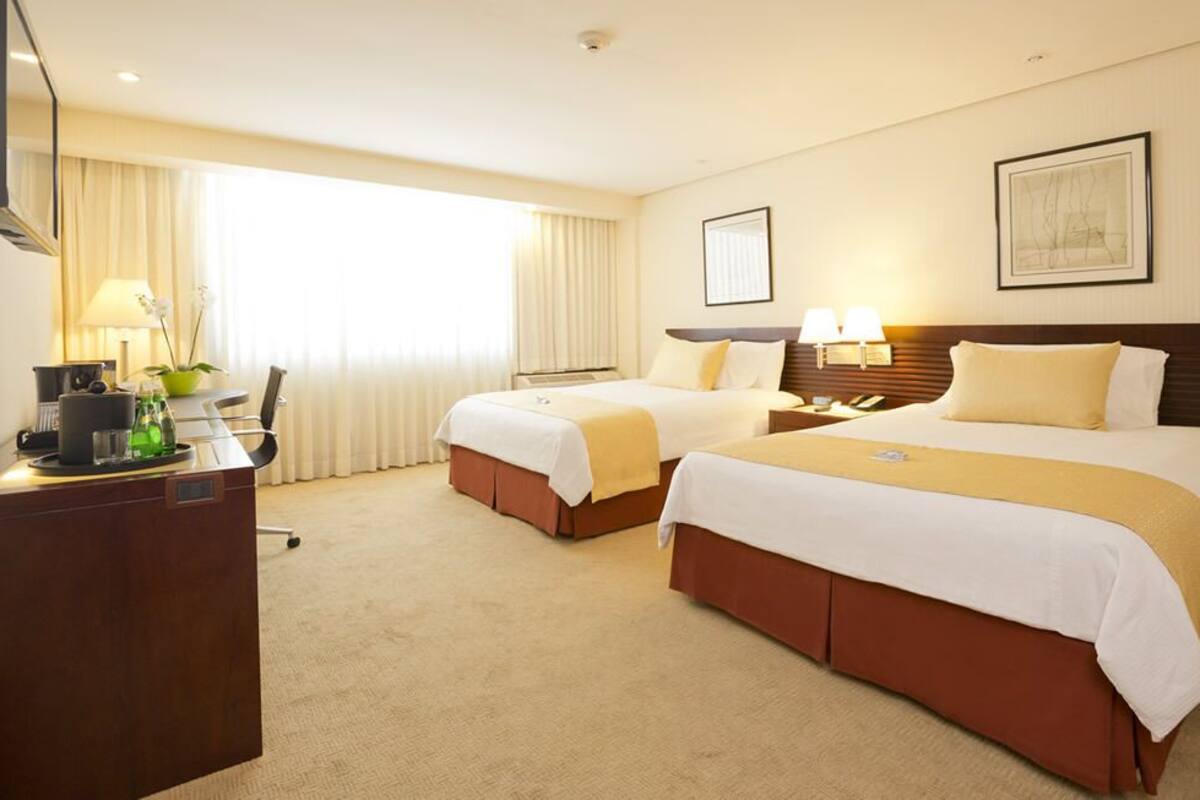 A hotel room with two beds, a desk, a chair, a lamp, and a window with curtains.