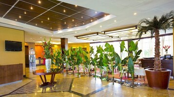 Um hall de hotel com teto azulejado, uma mesa de recepção e uma fileira de plantas em vasos.