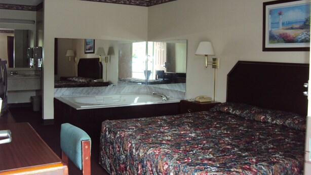 A hotel room with a bed, a desk, a chair, a bathroom with a sink and mirror, and a painting on the wall. - Western Motel (Folkston)