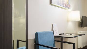 A hotel room with a blue chair, a desk with a lamp, a flat-screen TV, and a painting on the wall.