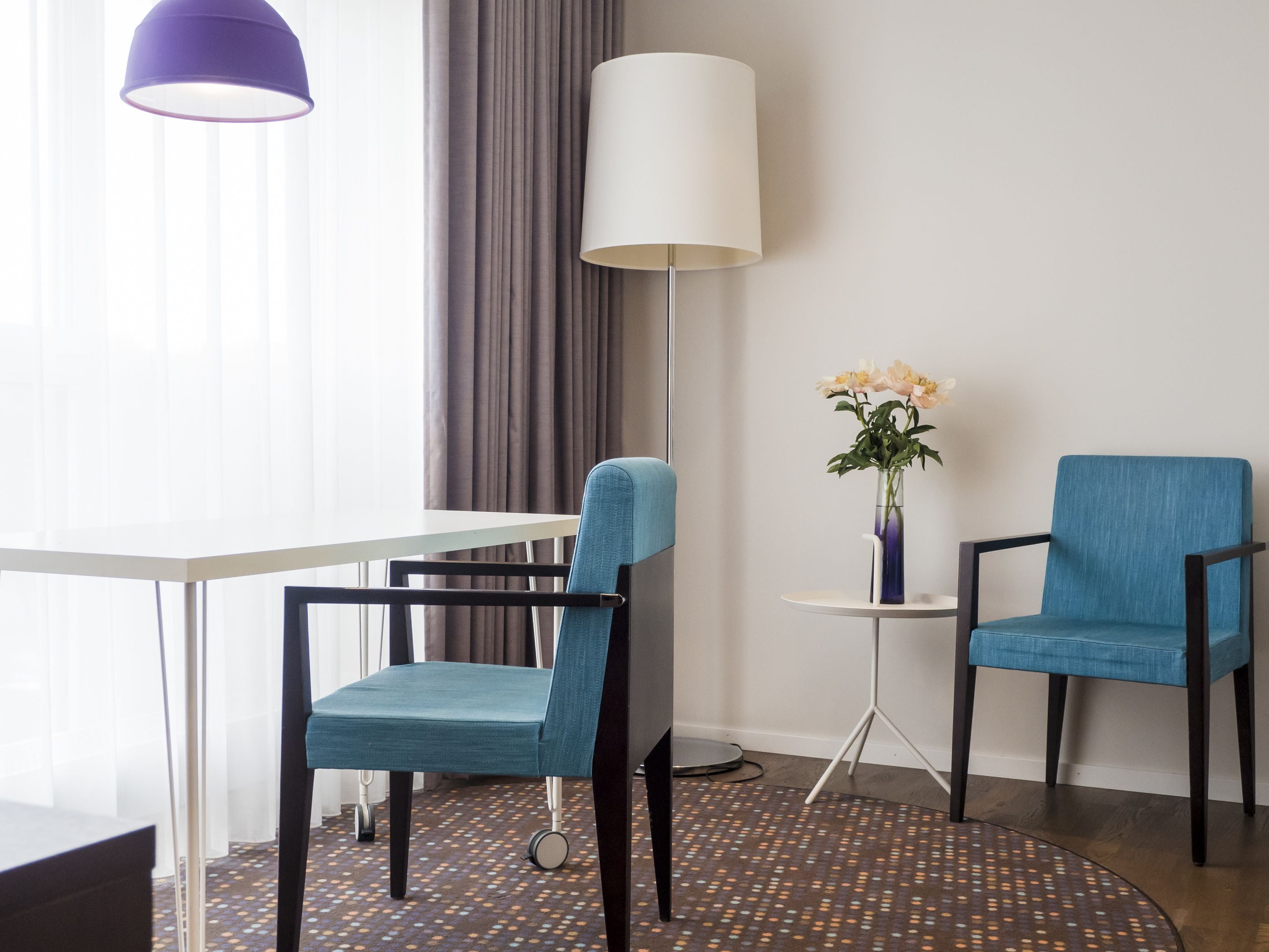 A hotel room with a white table, two blue chairs, a floor lamp, a vase with flowers, and a patterned carpet.