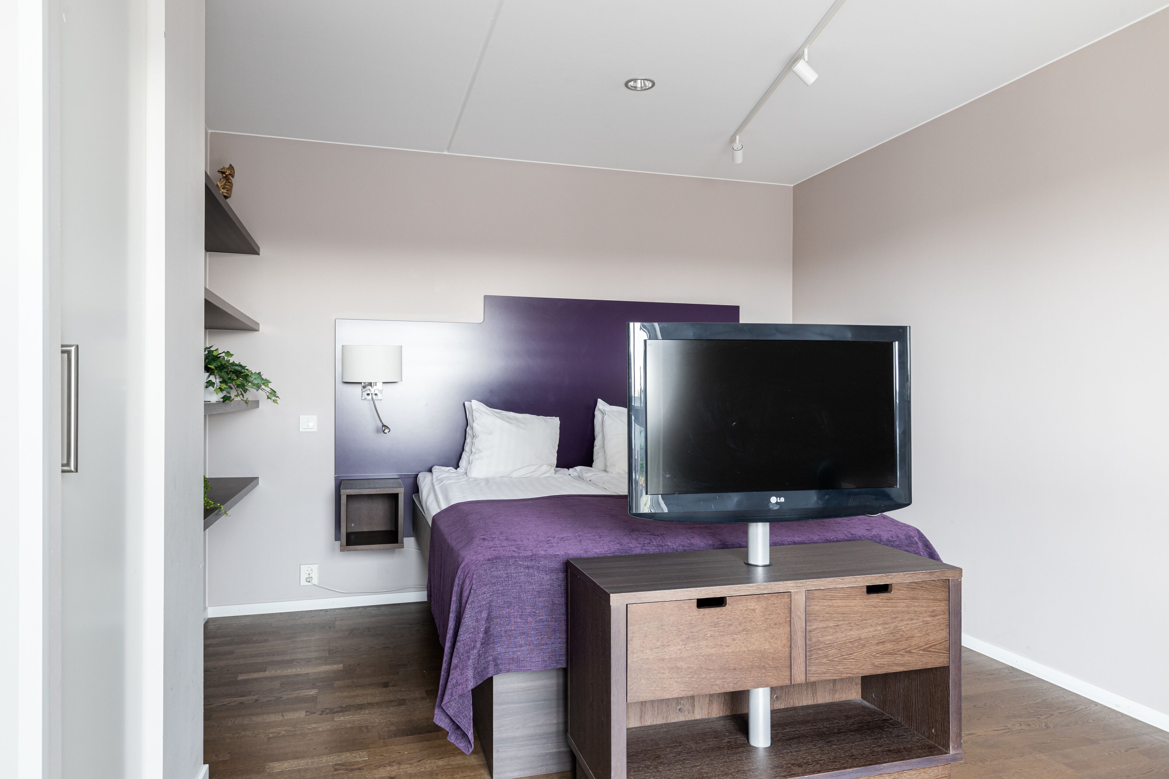 A modern bedroom with a bed, a TV on a wooden stand, a bedside table, and a wall-mounted shelf with a plant.