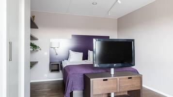A modern bedroom with a bed, a TV on a wooden stand, a bedside table, and a wall-mounted shelf with a plant.