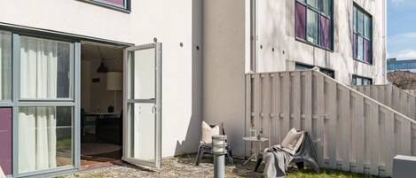 A modern building with a glass door entrance, a small outdoor seating area with chairs, and a wooden fence.