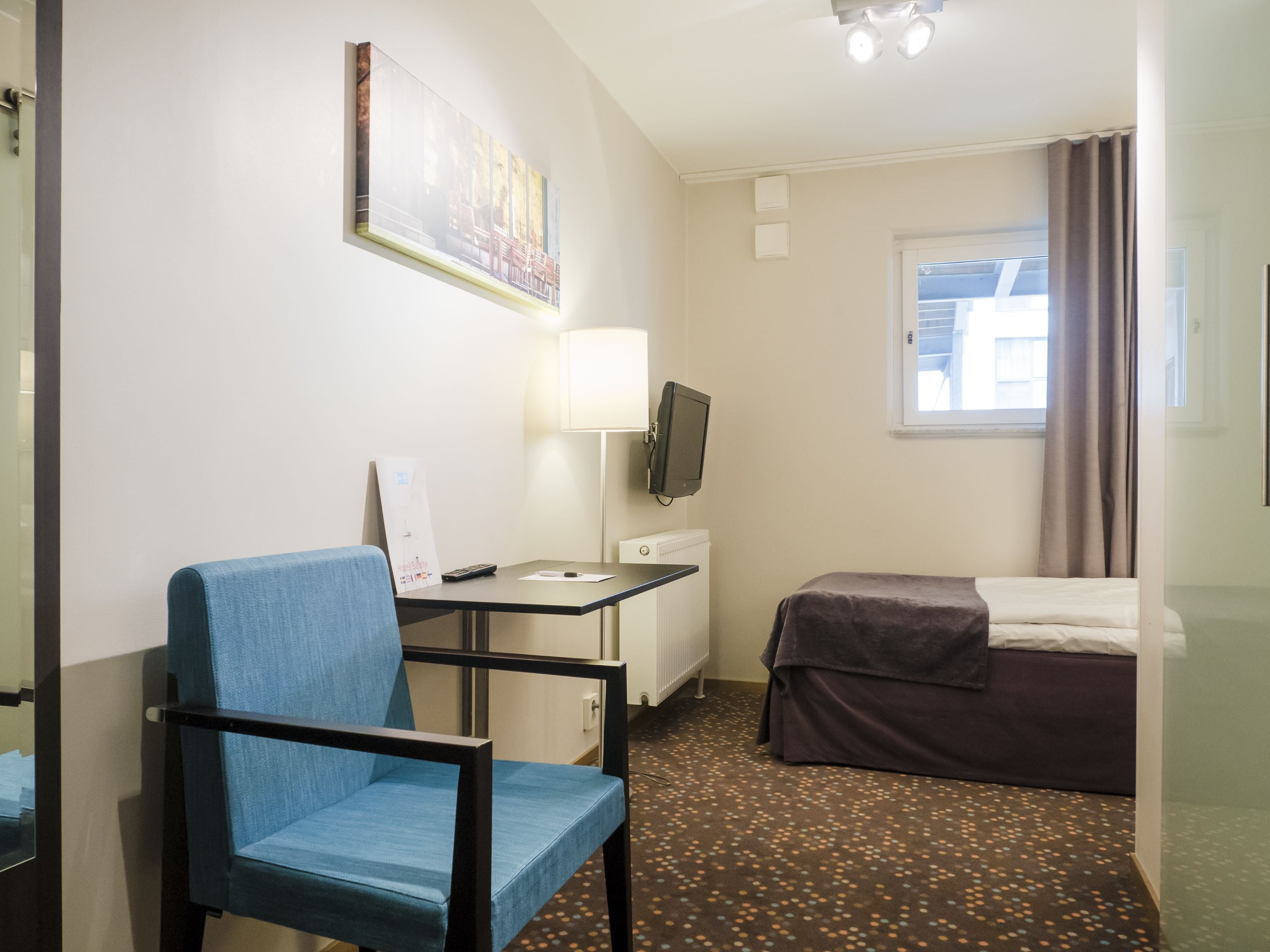 A hotel room with a single bed, a desk with a chair, a television, and a window with curtains.