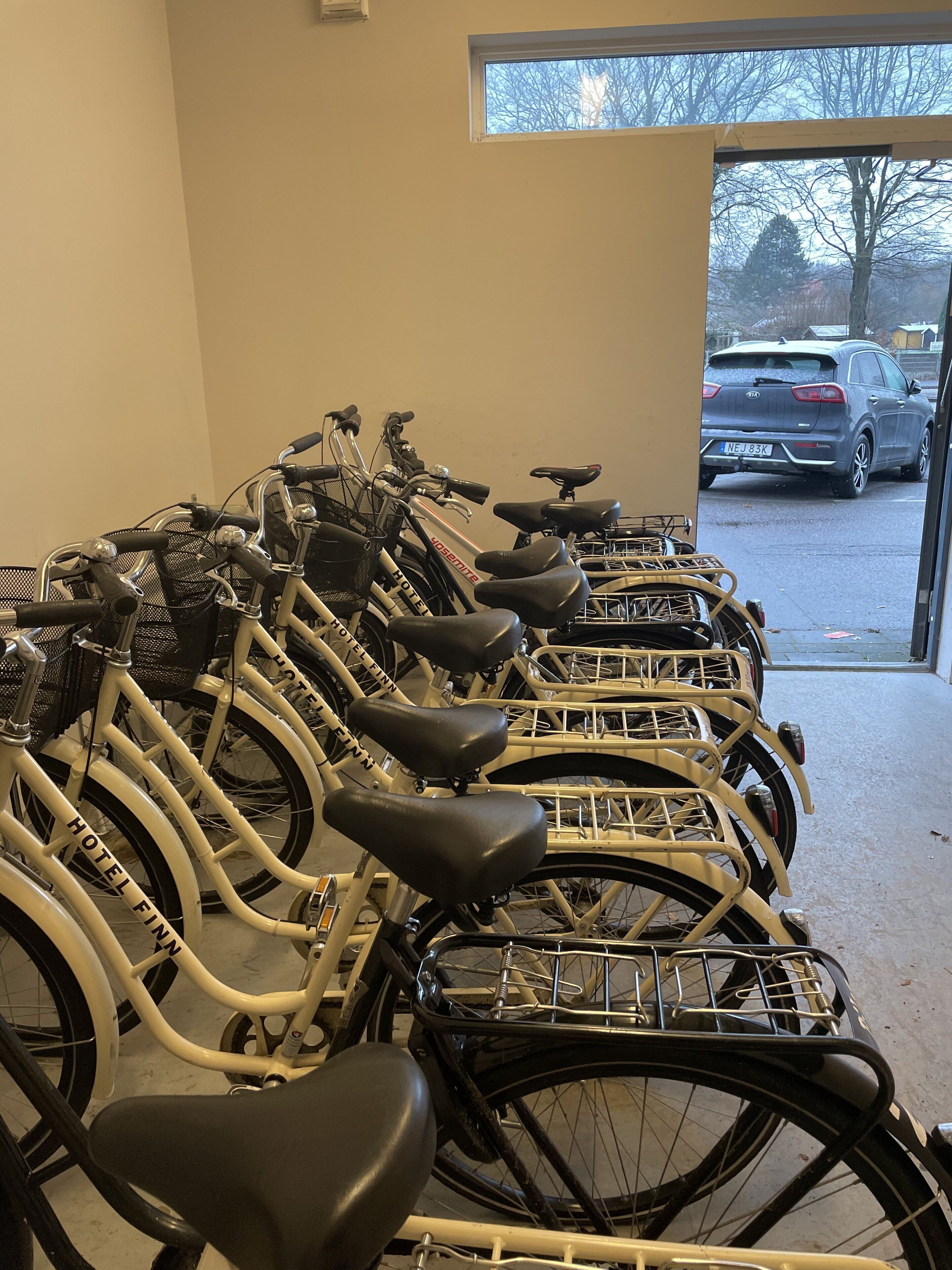 A row of bicycles with "HOTEL FINN" branding, parked indoors with a view of a parking lot outside.