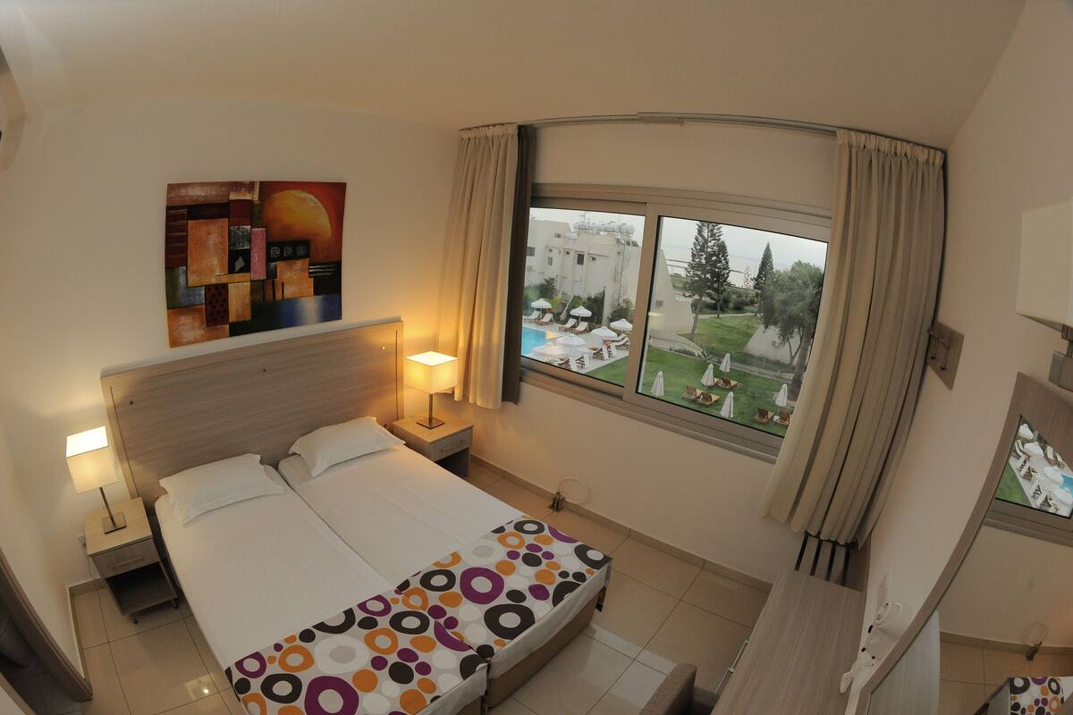 Quarto de hotel com cama, mesa de cabeceira, luminária, pintura na parede e janela com vista para uma piscina e vegetação.
