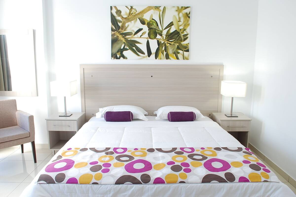 Quarto de hotel com uma cama, duas mesas de cabeceira, uma cadeira e uma obra de arte na parede.