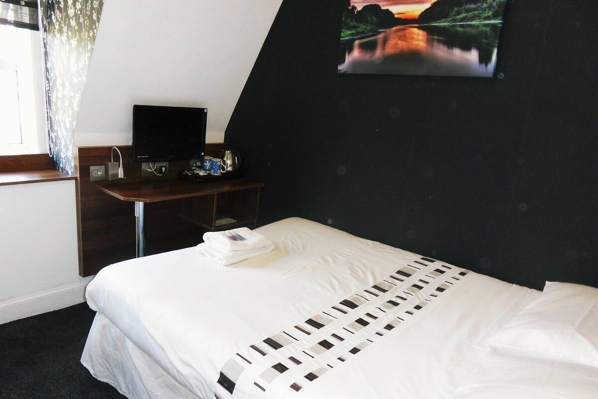 Ein Hotelzimmer mit einem Bett, einem Schreibtisch mit Computer und Kaffeemaschine, einem Bild an der Wand und einem Fenster mit Vorhängen.