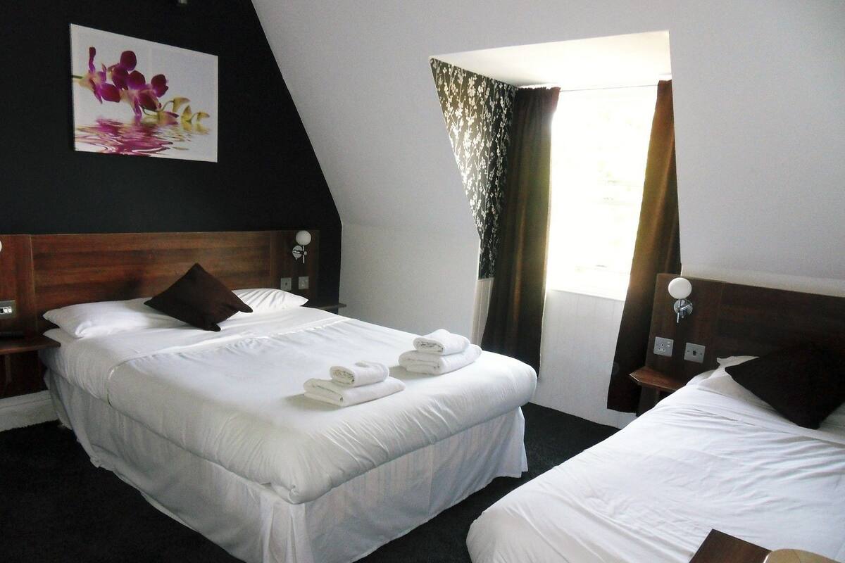 Ein Hotelzimmer mit zwei Betten, einem Holzkopfteil, einem Blumenbild, einem Fenster mit Vorhängen und einem Nachttisch mit Lampe.