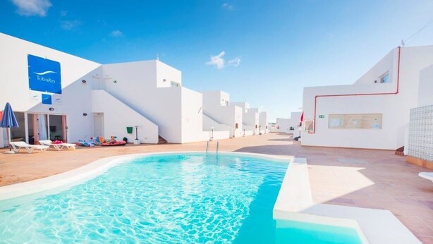 A pool area with white buildings, a blue sign, and a clear sky. - Tabaiba Apartamentos (Teguise)